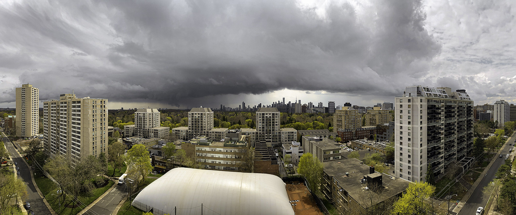 April 24, 2023 at 12:27 PM - Mid-spring view toward downtown Toronto just after noon: a dark storm shelf cloud sprawls overhead while budding green trees and high-rises sit beneath the dramatic sky