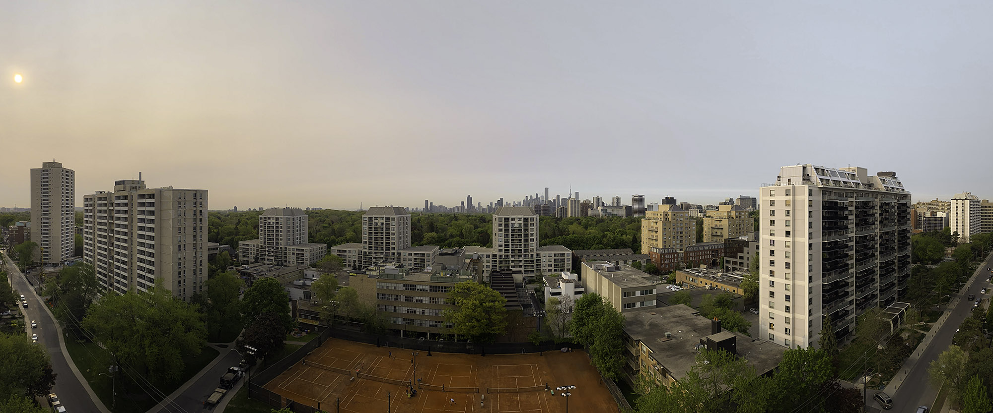 May 16, 2023 at 7:56 AM - Early-morning smoky haze over Midtown Toronto: muted sun on the left, newly leafed trees and clay tennis courts below, city skyline faint on the horizon