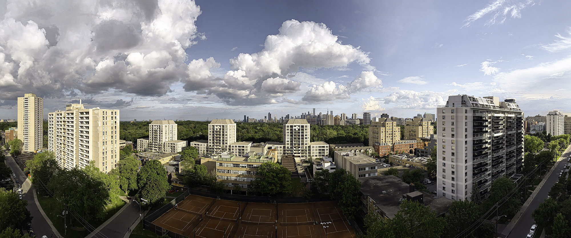 June 24, 2023 at 6:39 PM - Early-summer evening panorama: vivid blue sky dotted with towering white cumulus clouds, warm sunlight on lush green canopy, tennis courts, and high-rise apartments