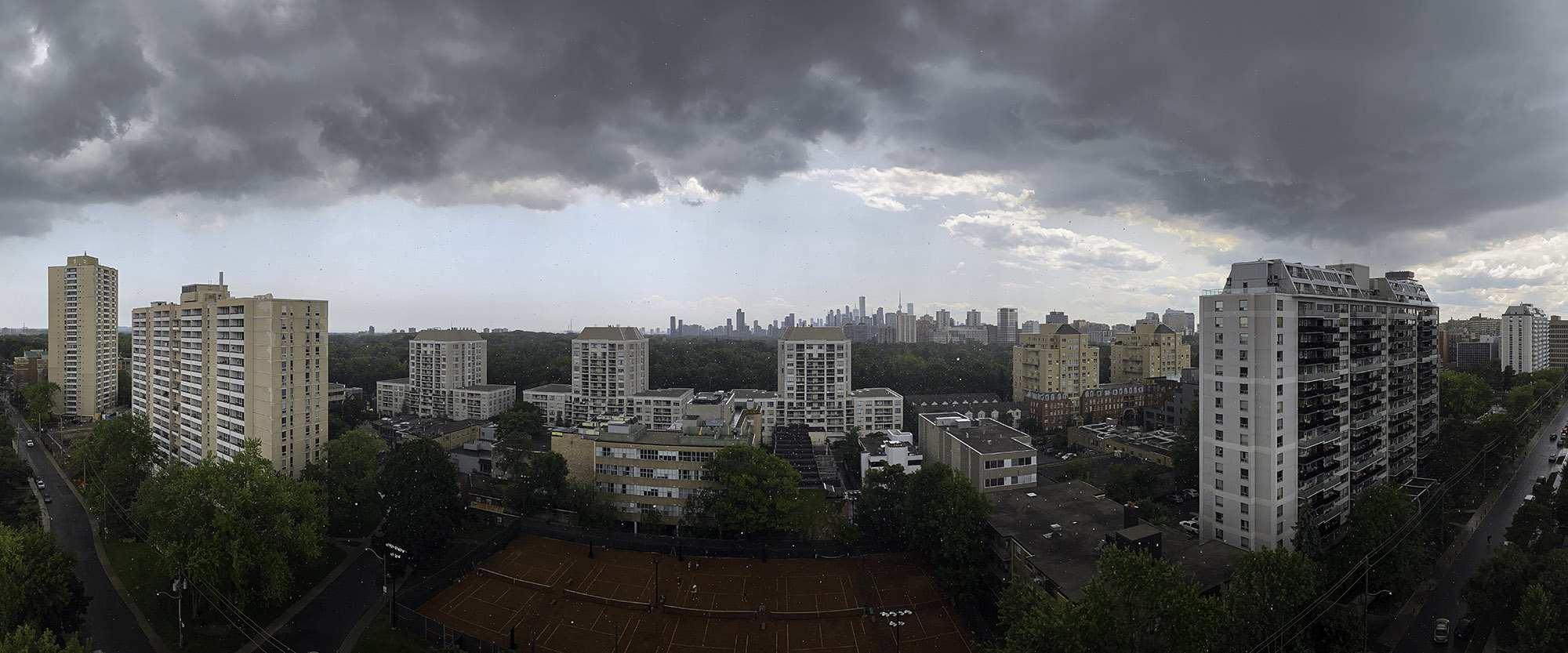 July 18, 2023 at 2:02 PM - Mid-afternoon thunderstorm rolling in: low, charcoal-gray clouds streak across the scene as light rain speckles the lens, contrasting with the green summer foliage and city towers