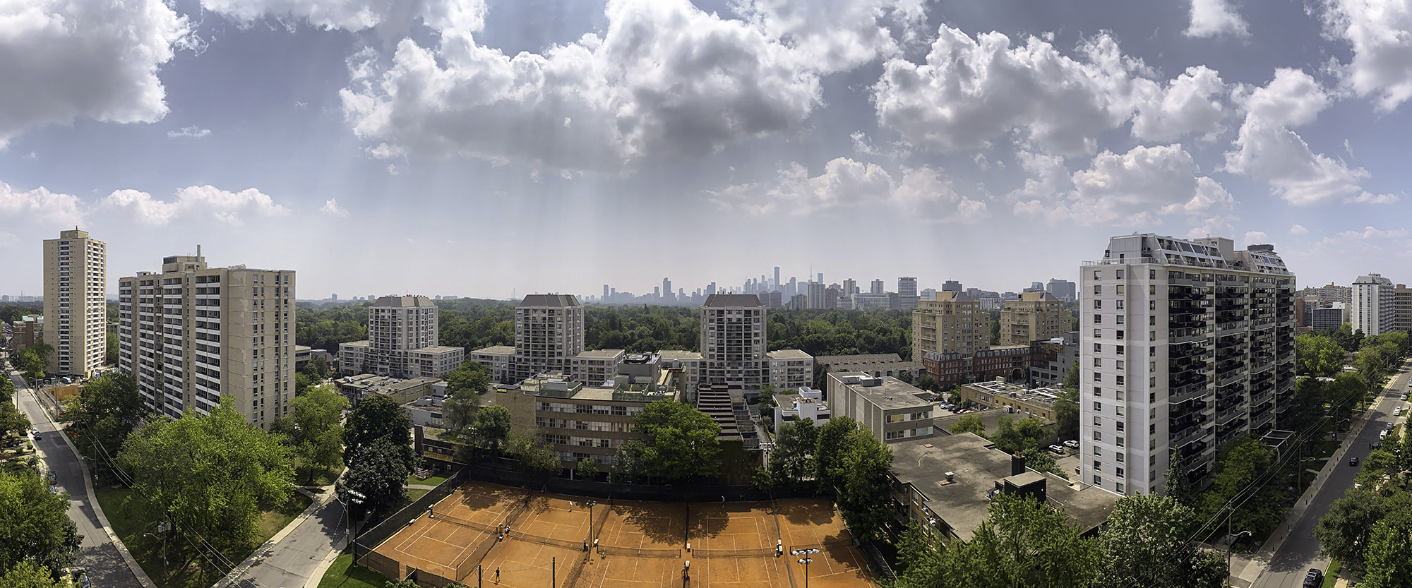 August 16, 2023 at 12:32 PM - High-summer midday scene: bright sunbeams break through scattered clouds, casting sharp shadows on vibrant green trees and busy orange clay courts beneath the Toronto skyline