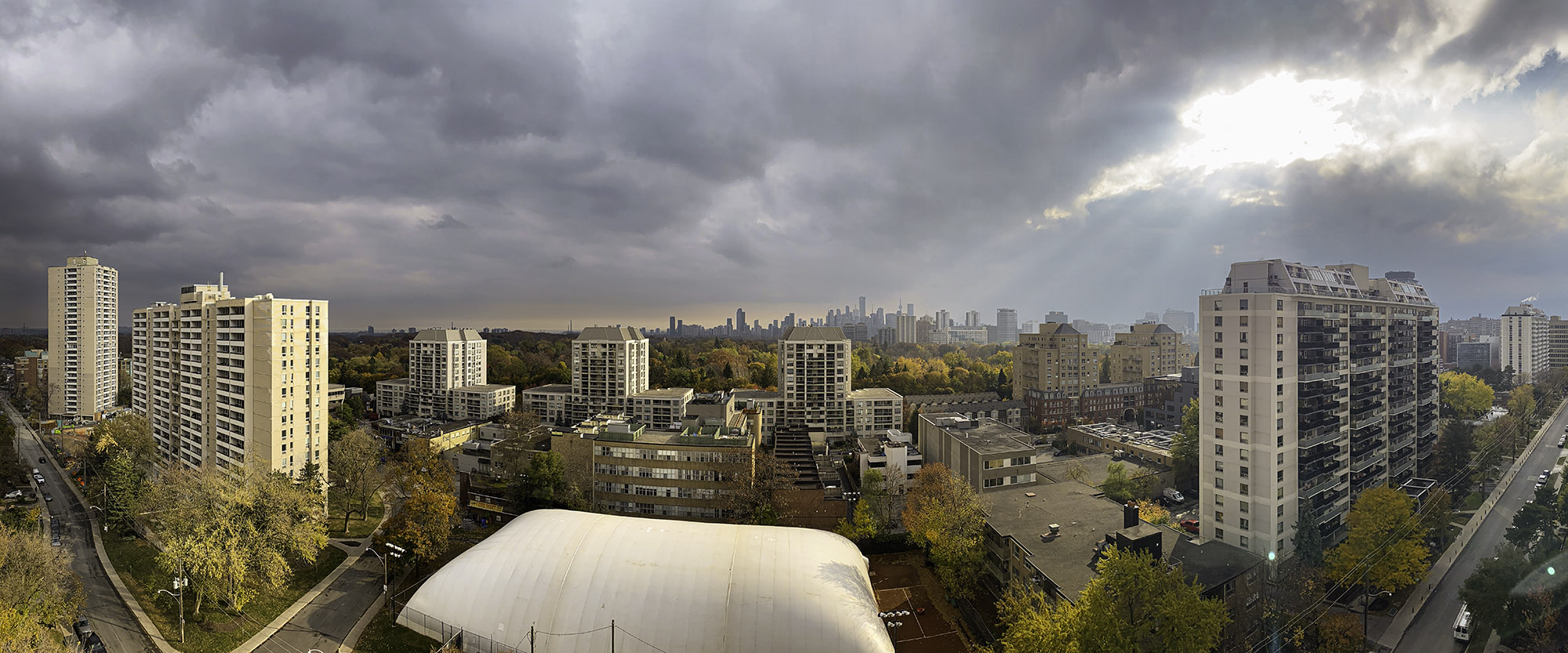 November 4, 2023 at 3:22 PM - Late-autumn afternoon: golden leaves and a white tennis dome glow under sun rays streaming through a break in heavy clouds, with downtown Toronto silhouetted in the distance