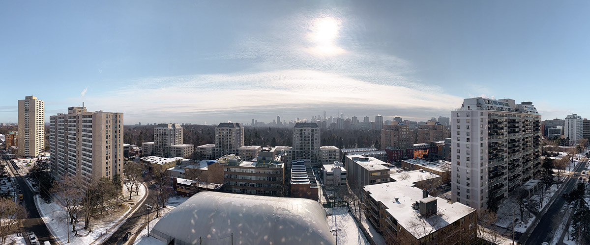 Open blue sky with a thin veil of high cloud around the sun, snow lying across rooftops, streets and the dome below.