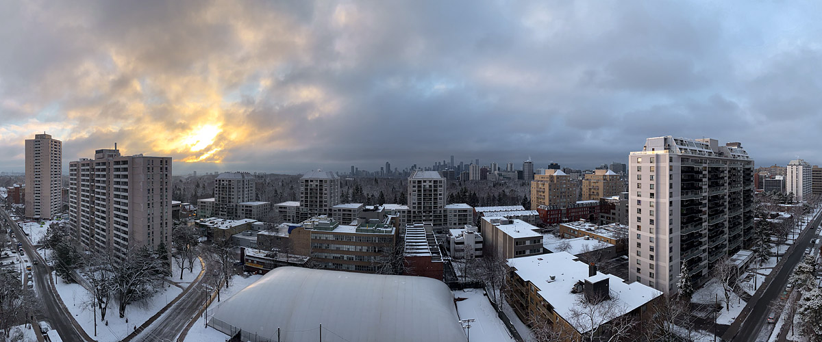 A dark grey cloud deck sits low over the skyline with a warm break of light to the left, snow covering rooftops and streets.
