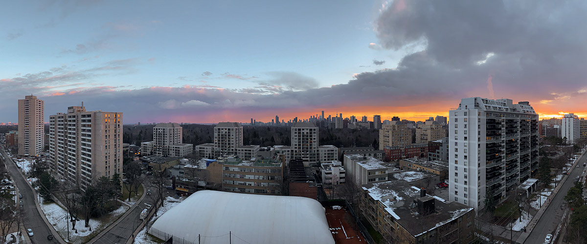 Orange light runs along the horizon under pink and grey cloud, with patches of snow on the roofs and grass below.