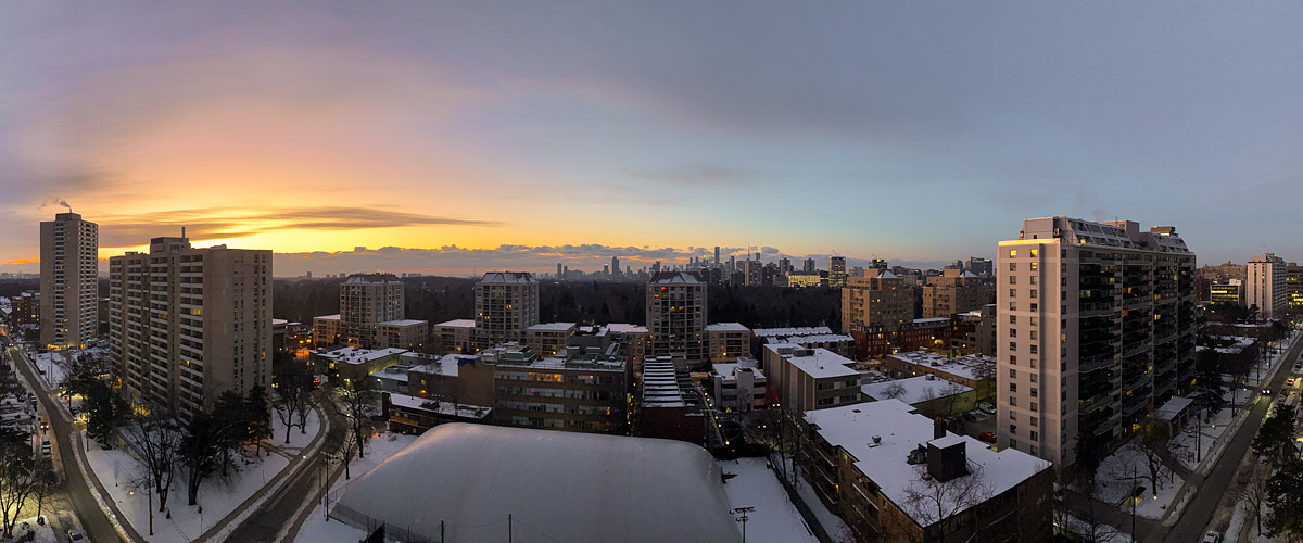 Deep orange and mauve fill the sky before sunrise, the skyline in silhouette and windows lit, snow across the ground.