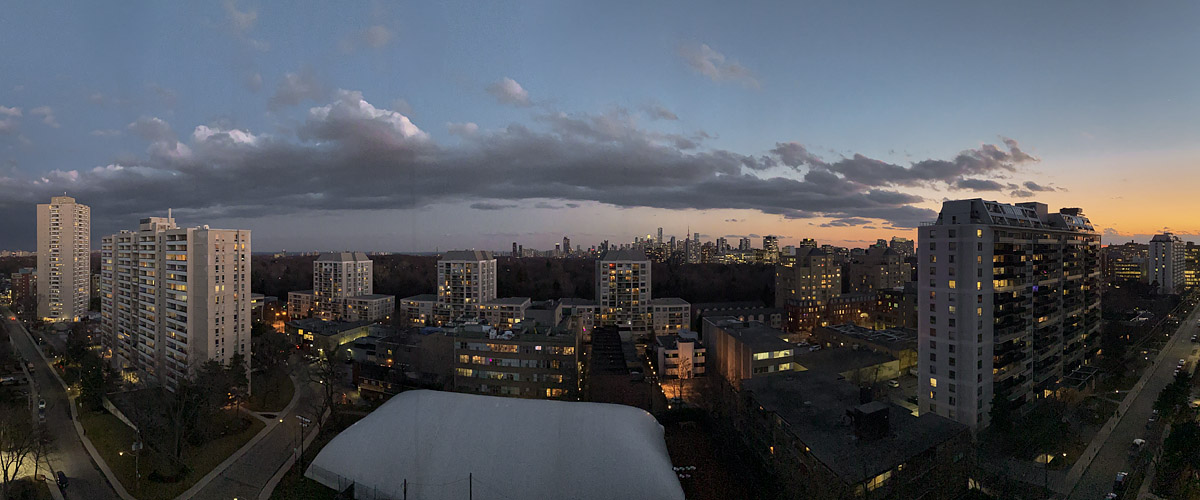 A dark broken cloud bank lies across the sky with a band of orange at the right horizon, the city lights on below.