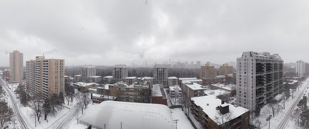 Light snow falls through flat grey air, the skyline faint behind it and the rooftops and courts covered in white.
