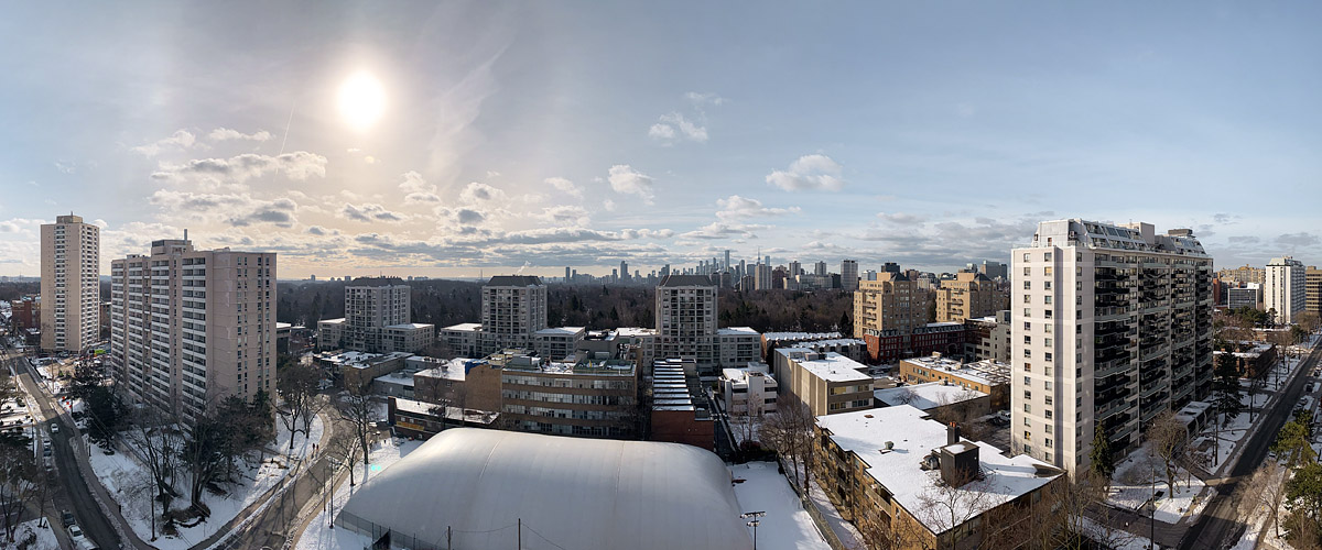 A bright sun in thin high cloud on the left, small clouds strung along the horizon under open blue, snow lying across the ground.