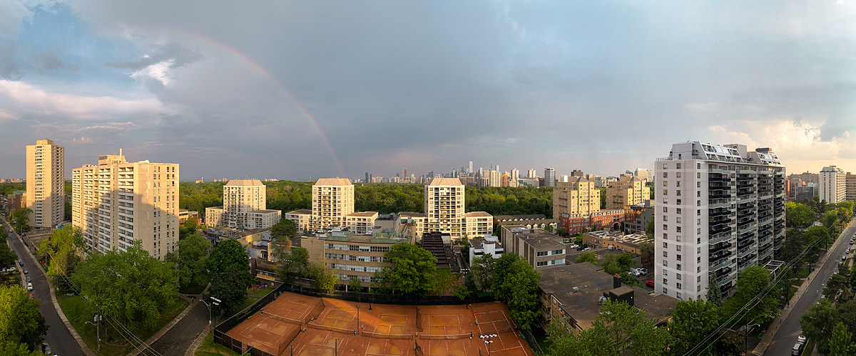 A rainbow arcs through grey rain cloud over the skyline while low sun lights the clay courts and treetops.