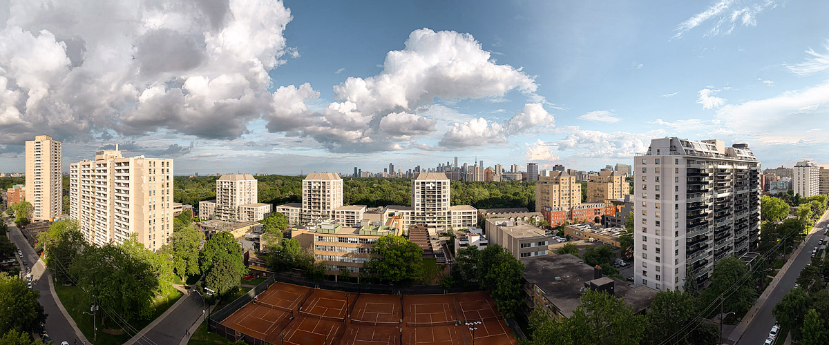 Towering white cumulus fill a blue sky, warm sunlight across the green canopy and clay courts.