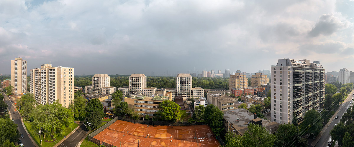 A grey cloud deck with a dark curtain of rain over the downtown towers, warm light reaching the courts.