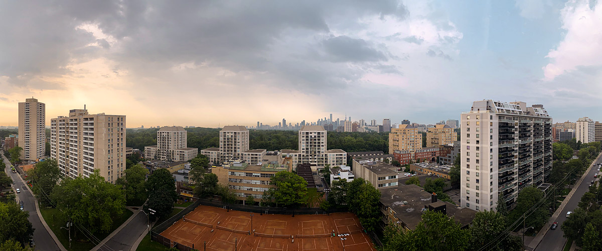 A grey cloud deck with a warm break on the left, rain trailing over the city and the skyline soft.