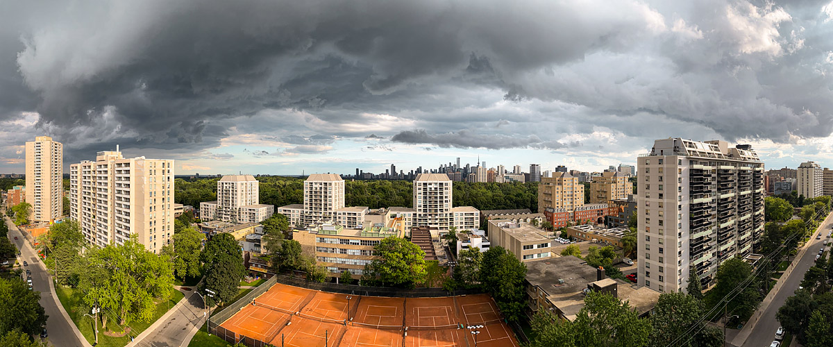 Dark storm cloud with a ragged underside covers the frame, a bright band of light along the horizon and sun on the courts.