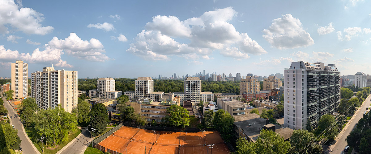 Bright white cumulus scattered across a blue sky, the skyline hazy behind them and warm light on the tower faces and the clay courts.