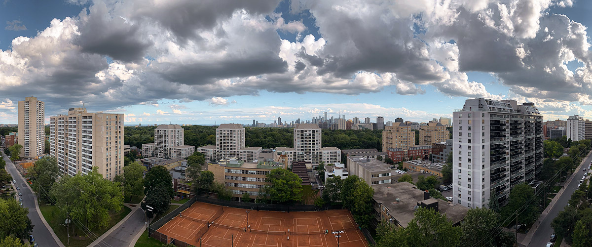 Large white and grey cumulus with blue between, the skyline clear beyond the trees and the clay courts open below.
