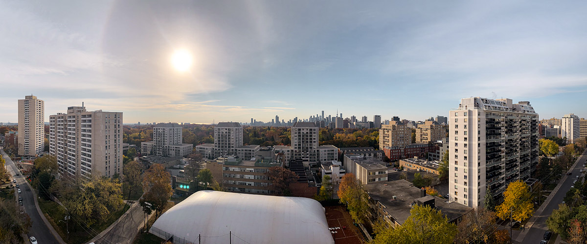 Thin high cloud with a solar halo, the city pale and low in contrast behind it.