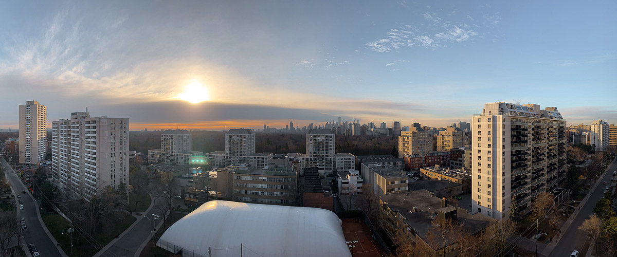 The low sun glows behind the edge of a cloud layer with pale sky above and a warm horizon behind the skyline.