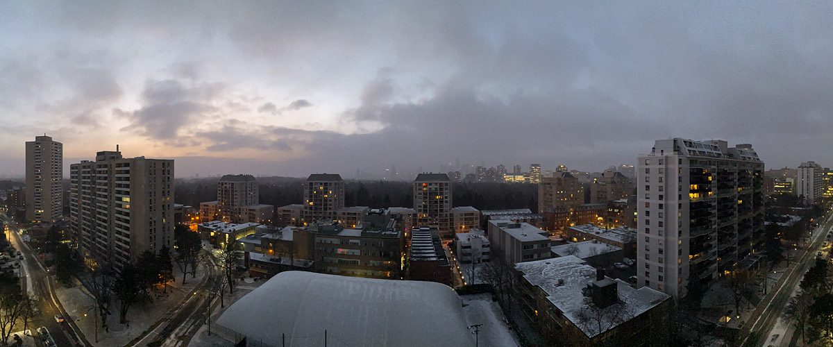 Heavy grey cloud with a faint warm break, fresh snow across the roofs and streets and the lights on.