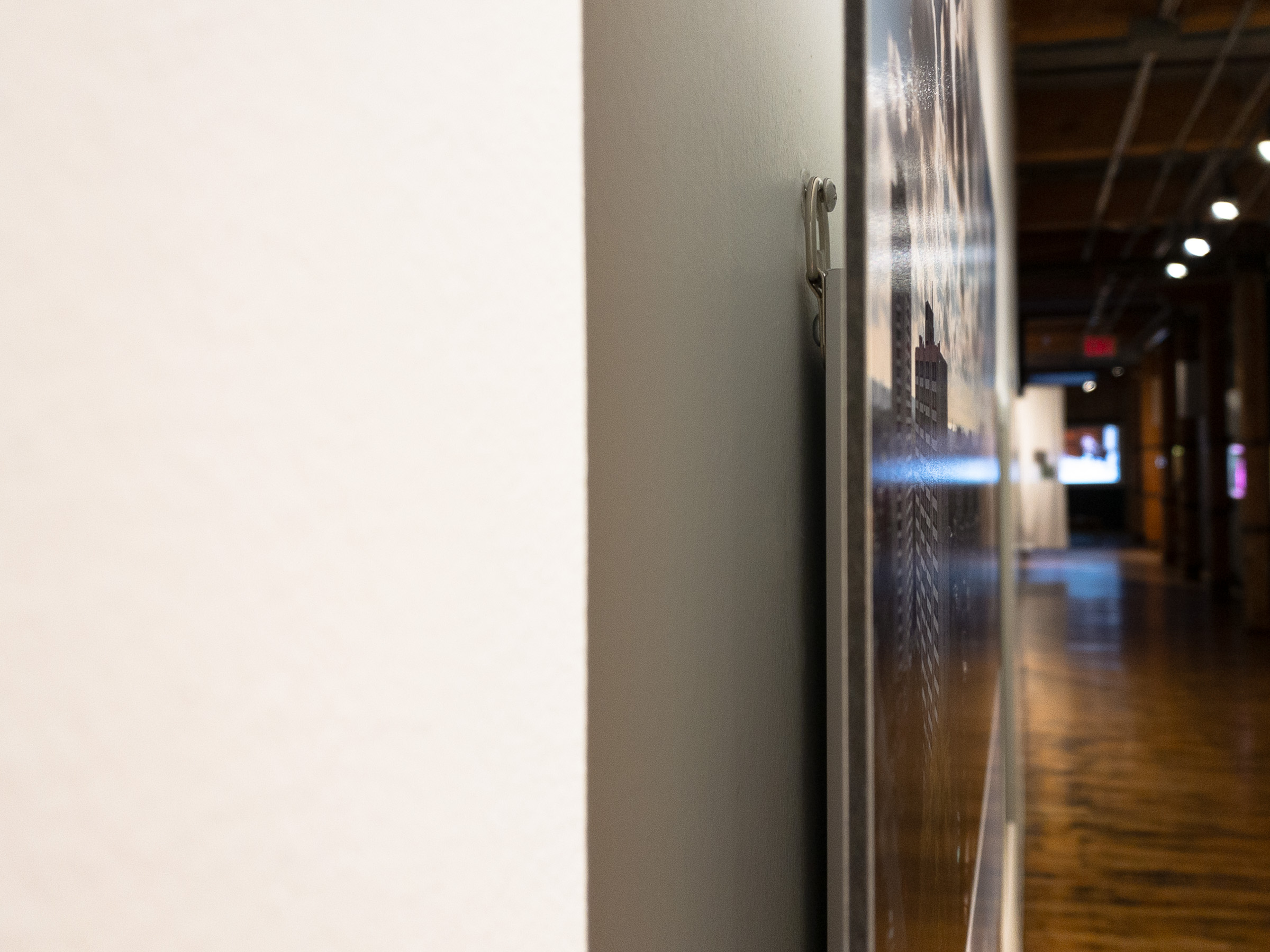 Side view of a mounted panel: its edge, the hanging hardware, the gap and shadow against the wall, and the corridor beyond.